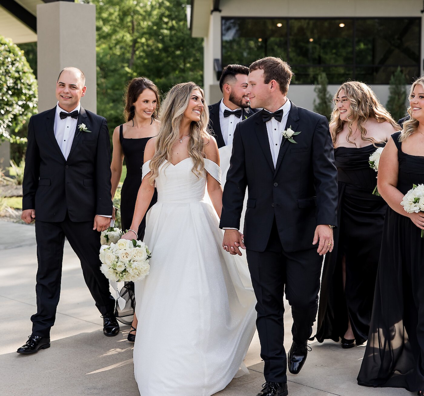 Joyful wedding party portrait of the bride and groom leading their attendants while walking and holding hands. The wedding party wears a classic color scheme: the men in black tuxedos and the women in various styles of black dresses.