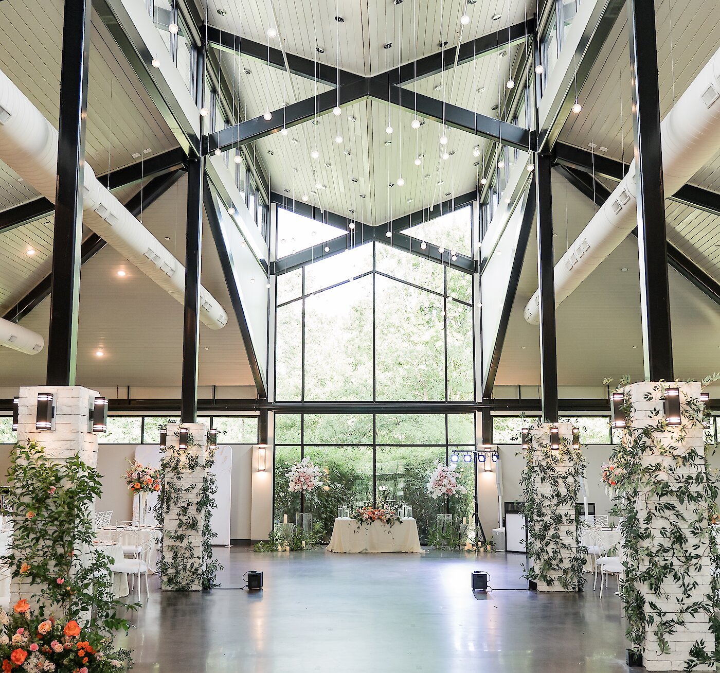 Modern wedding ceremony space featuring tall glass windows, black steel beams, and lush floral arrangements lining white stone pillars, leading toward a beautifully decorated sweetheart table.