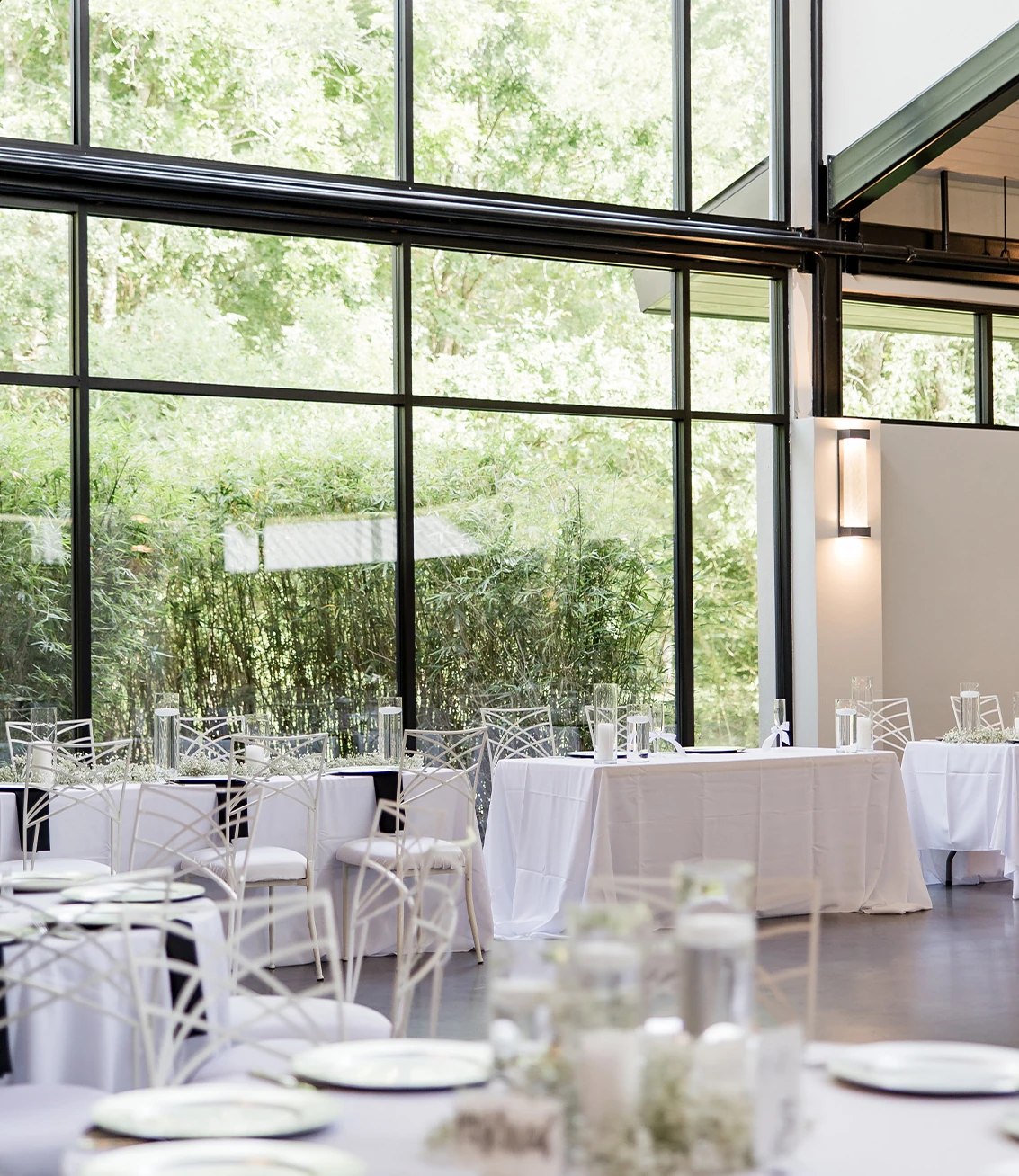 A reception table setting featuring a gold-stand centerpiece with abundant greenery and small white flowers, gold charger plates, and white chairs in a vaulted ballroom.