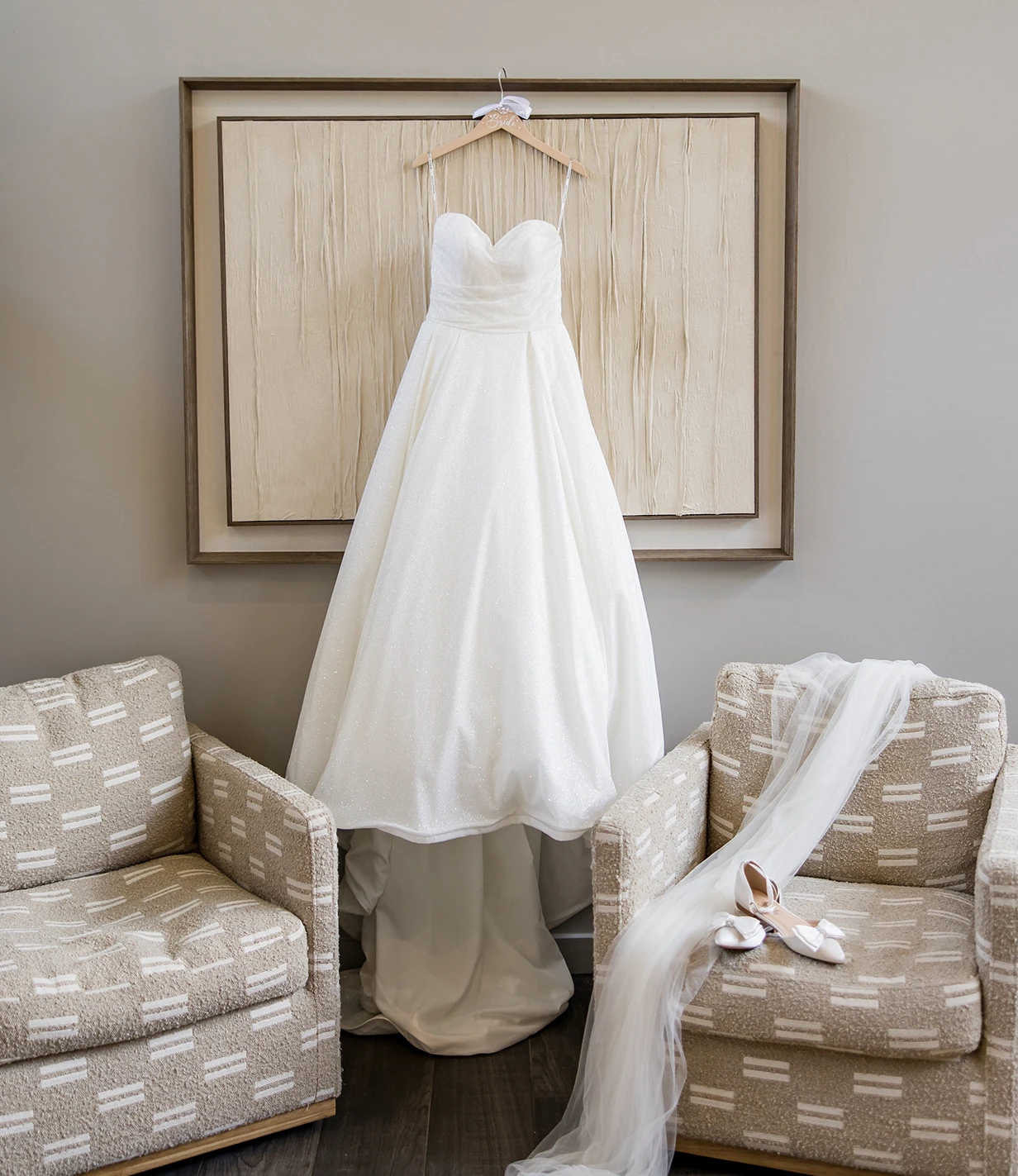 White wedding dress hanging on a wooden frame between two upholstered chairs, with the bride’s veil and shoes draped over one chair.