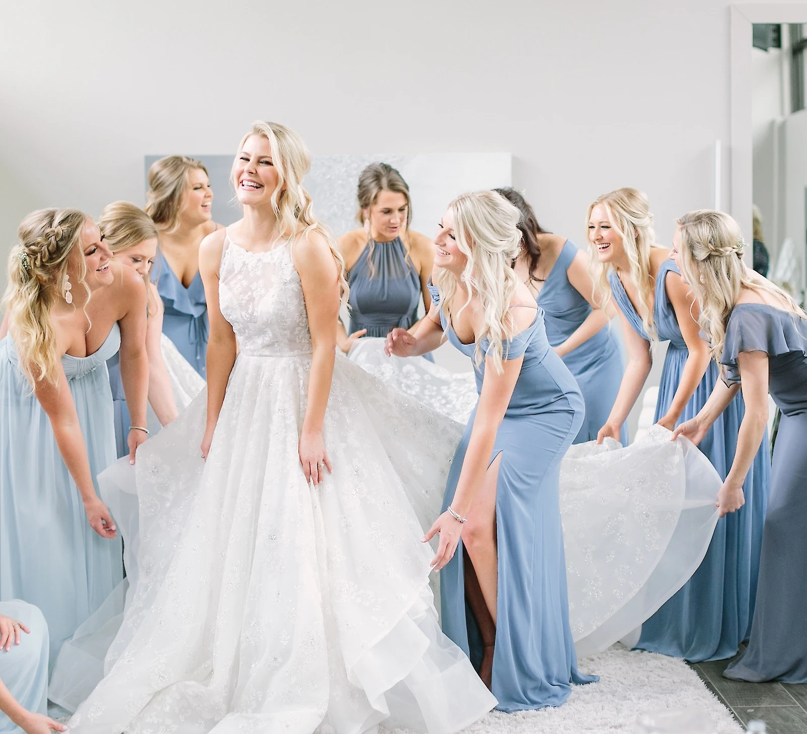 The laughing bride stands in her white lace gown while seven bridesmaids in varying styles of dusty blue dresses fluff and hold out her skirt in the bright suite.