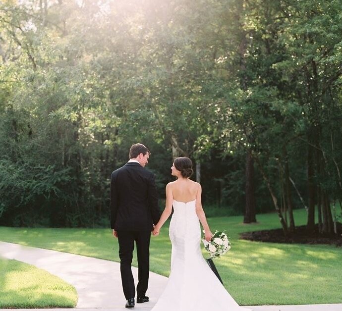 The bride and groom walk hand-in-hand away from the camera down a sunny paved path, looking at each other, surrounded by lush green trees.