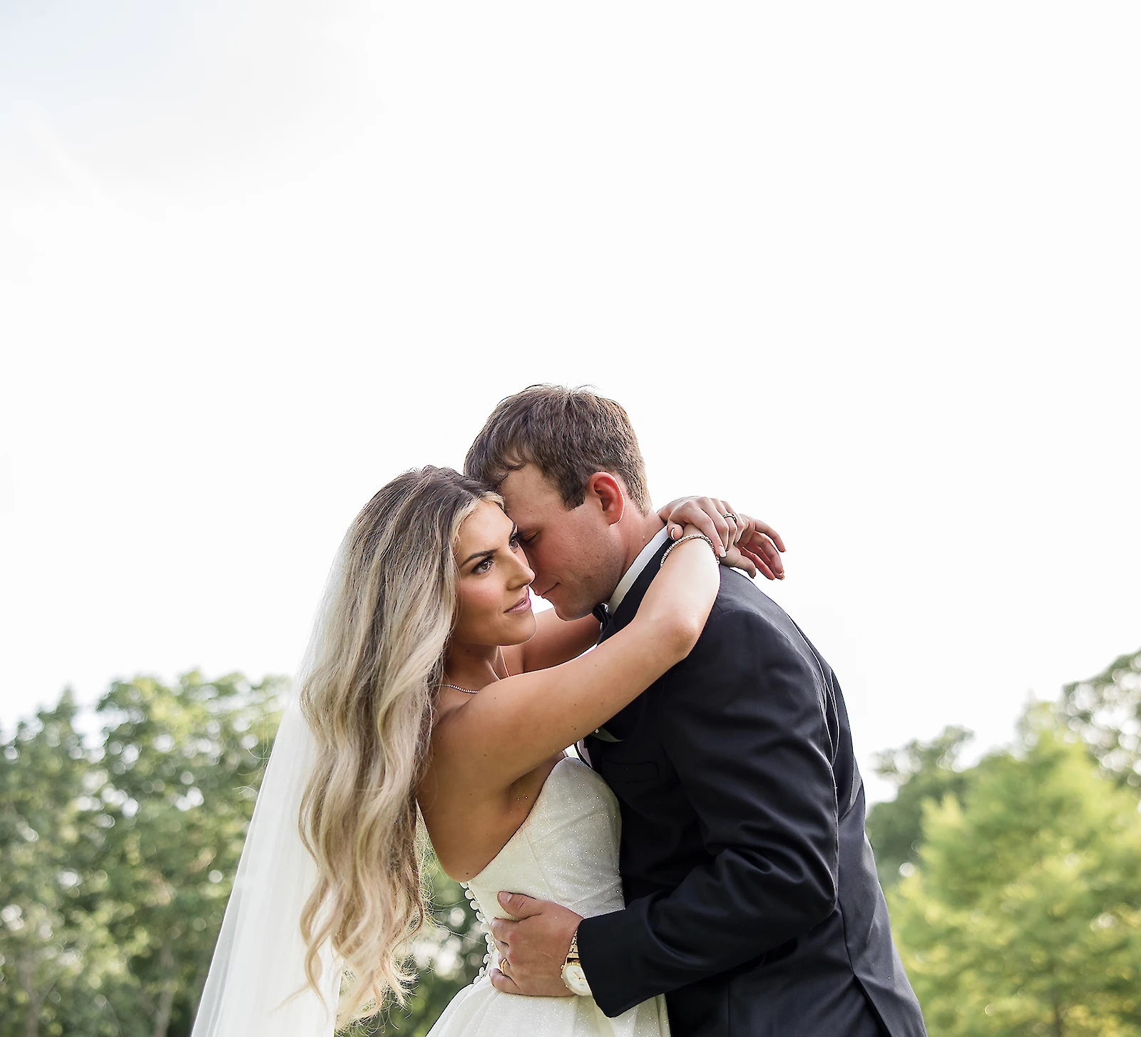 Bride and groom embracing outdoors during their wedding, with the bride’s veil flowing and trees in the background