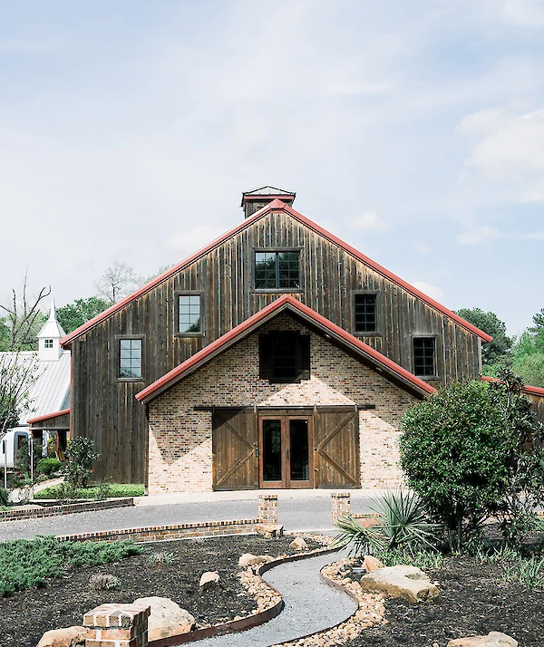 Rustic barn-style wedding venue with wood siding, brick accents, red trim, and a landscaped walkway leading to double wooden doors