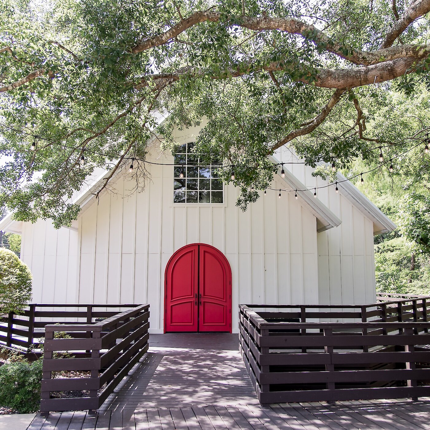 White chapel with bold red double doors, surrounded by lush green trees and hanging string lights, with a wooden walkway leading to the entrance.