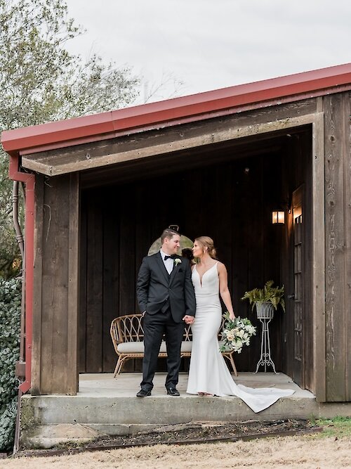 Bride and groom stand together in a rustic wooden shed with a red roof, holding hands and smiling, with the bride in a sleek white gown and the groom in a black tuxedo.
