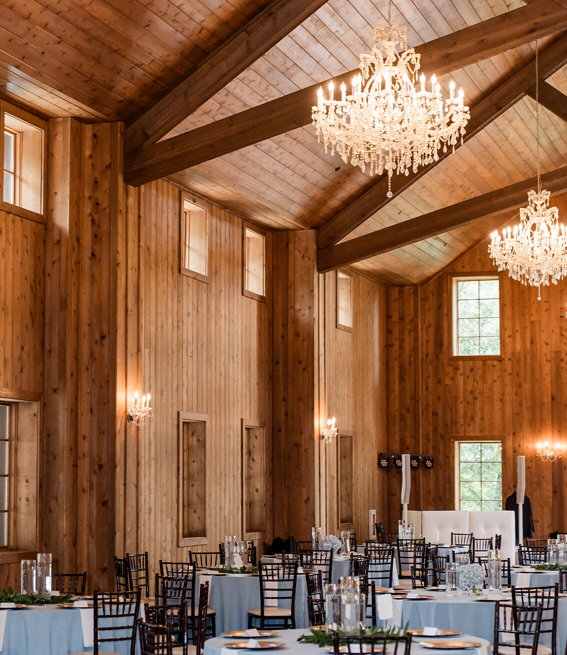 Elegant wedding reception hall with round tables set with white linens, gold chargers, floral centerpieces, and crystal chandeliers hanging from draped wooden ceiling beams.