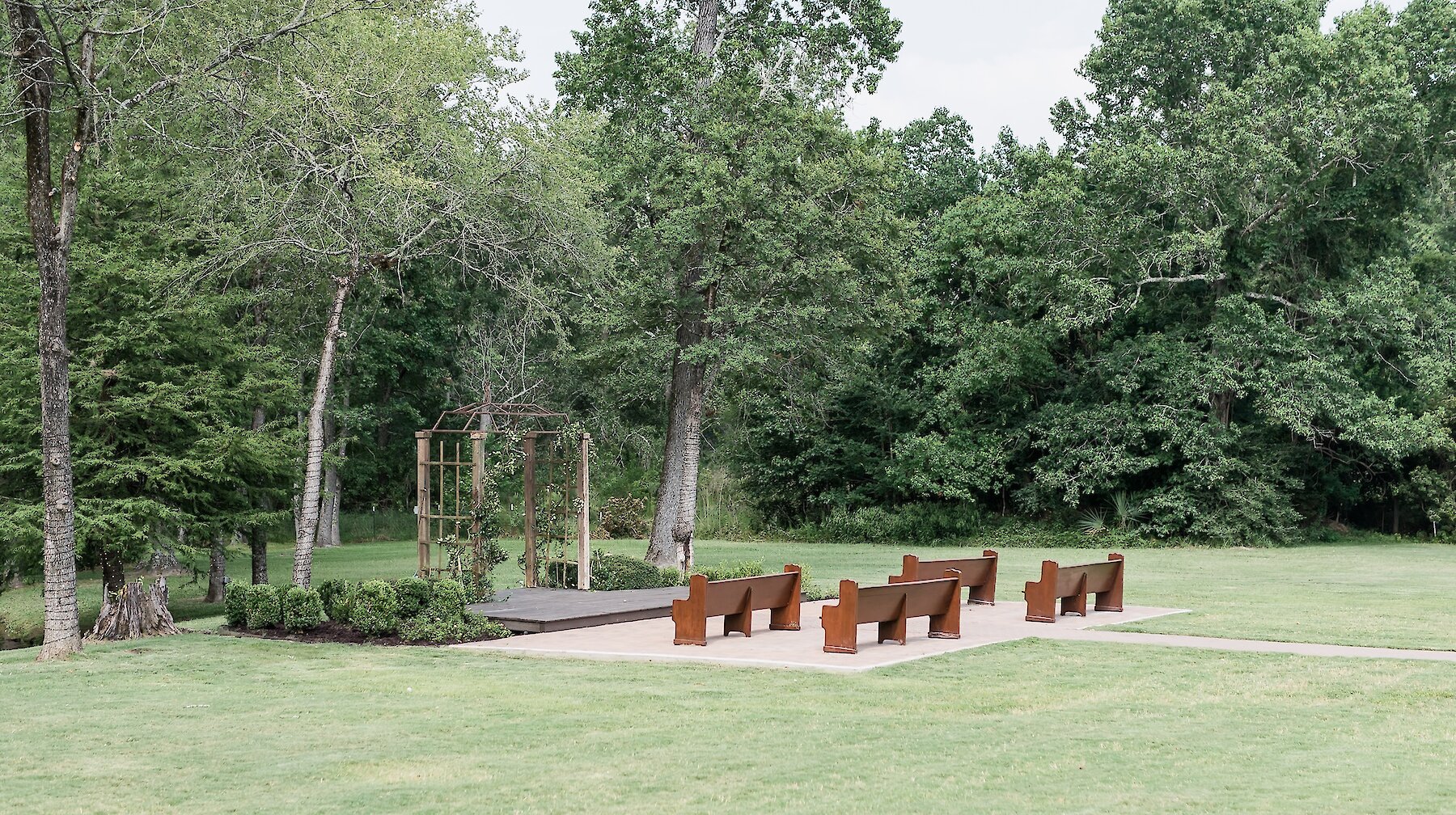 Outdoor ceremony space with wooden benches facing a small arbor surrounded by lush green trees and grass.