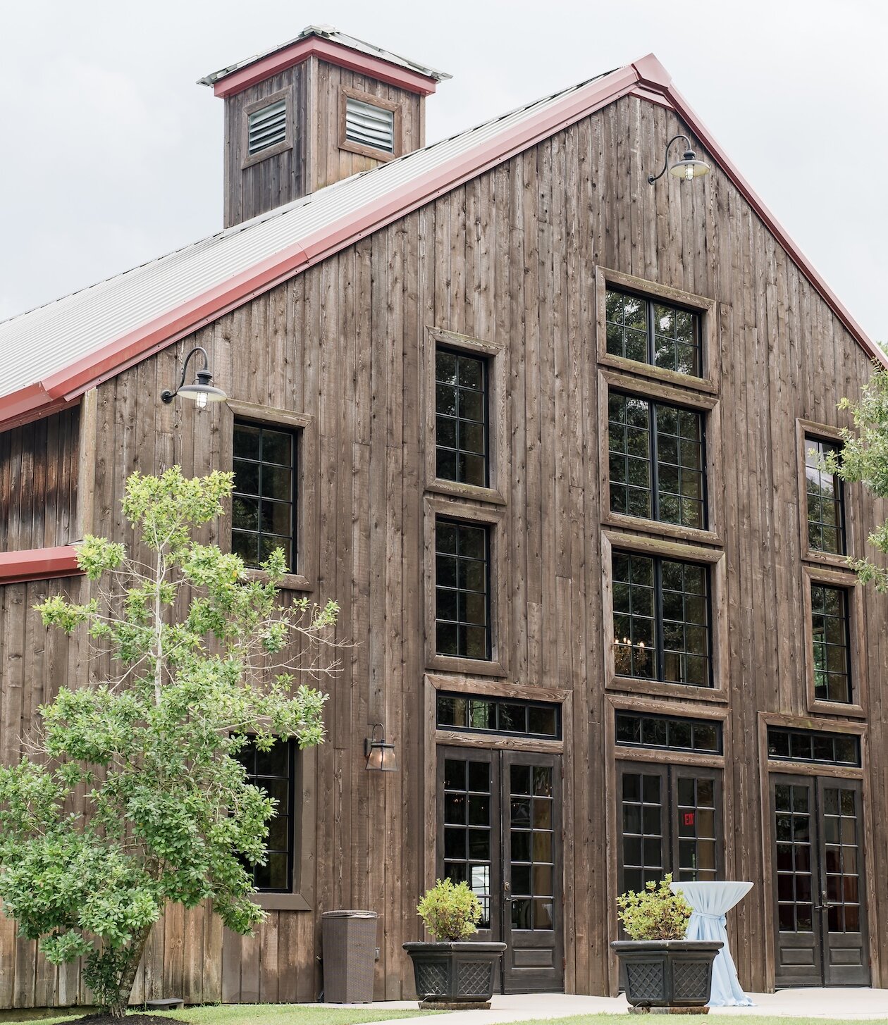 Large rustic wooden barn venue with tall black-framed windows, red roof accents, and cocktail tables draped in blue linens outdoors