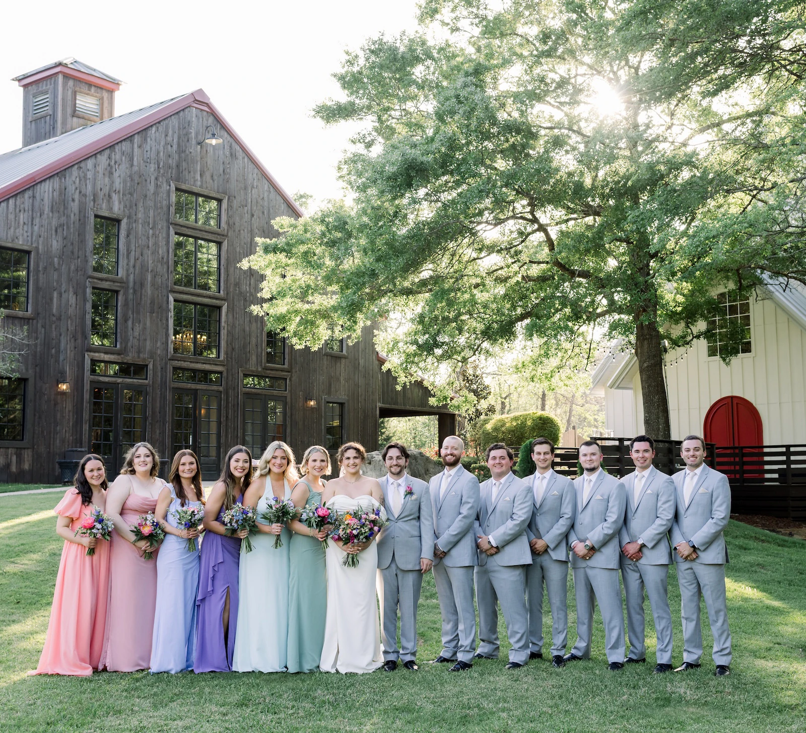 Wedding party portrait outdoors, featuring bridesmaids in pastel dresses and groomsmen in light gray suits standing beside the bride and groom in front of a rustic wooden barn and lush green trees.