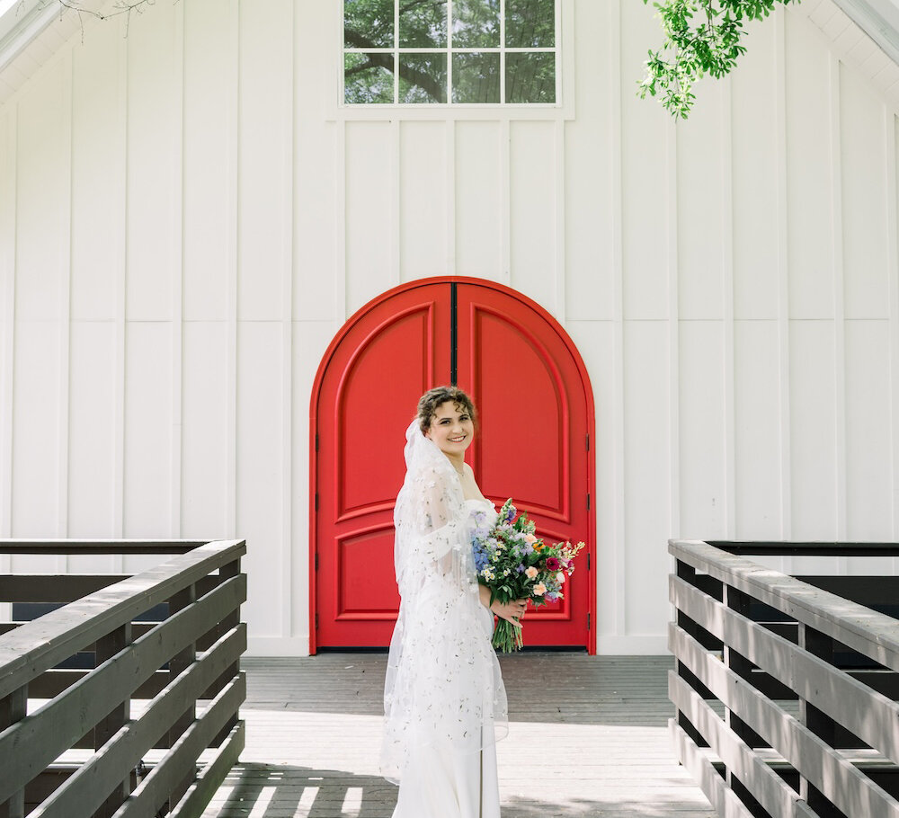 Bride stands on a wooden walkway in front of a white chapel with a bright red arched door, holding a colorful bouquet and smiling toward the camera.