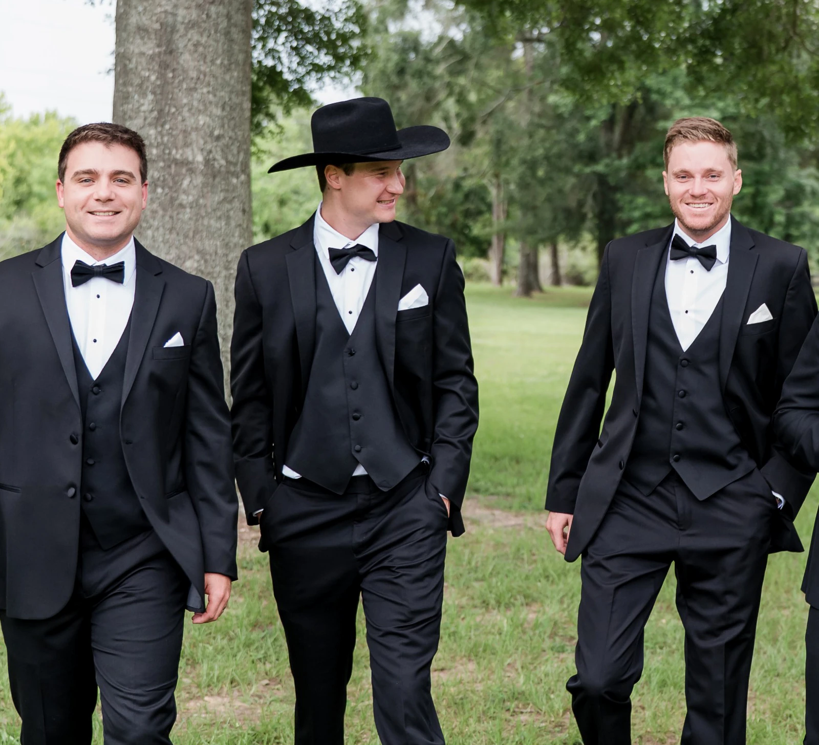Four groomsmen in black tuxedos walk together outdoors under a large tree, smiling and chatting in a grassy field.