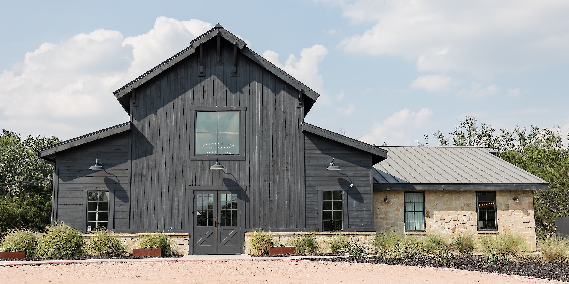 Modern black barn wedding venue exterior with vertical wood siding and a central gable, connected to a section built with rustic light-colored stone, standing under a bright blue sky.