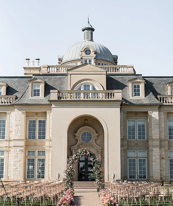 European-style building entrance with a tiered marble fountain and formal gardens at dusk.