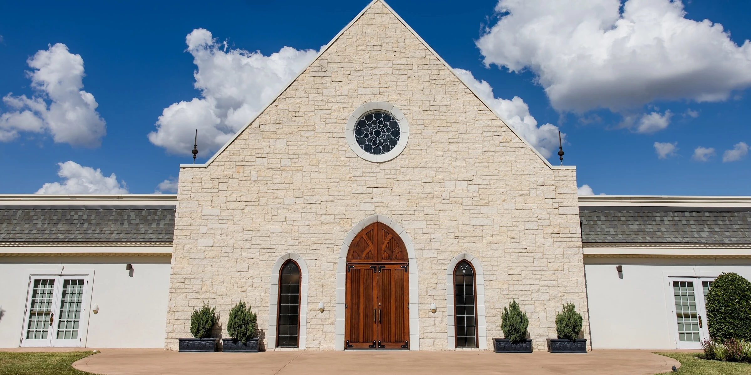 Stone chapel exterior with arched wooden doors, tall narrow windows, and a bright blue sky with scattered white clouds above.