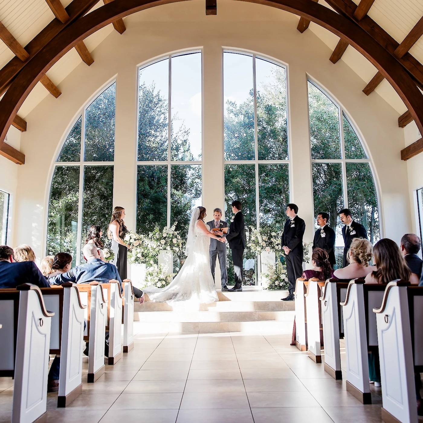 Bright wedding ceremony inside a vaulted chapel with exposed dark wooden arches and a dramatic wall of tall arched windows overlooking a forest. The couple is exchanging vows on a raised altar, flanked by the wedding party and guests seated in white pews.