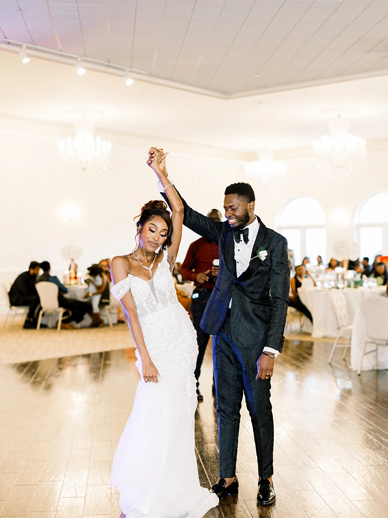 Bride and groom share a kiss on an elevated platform surrounded by tall windows and lush greenery, with pink floral vines framing the ceremony space.