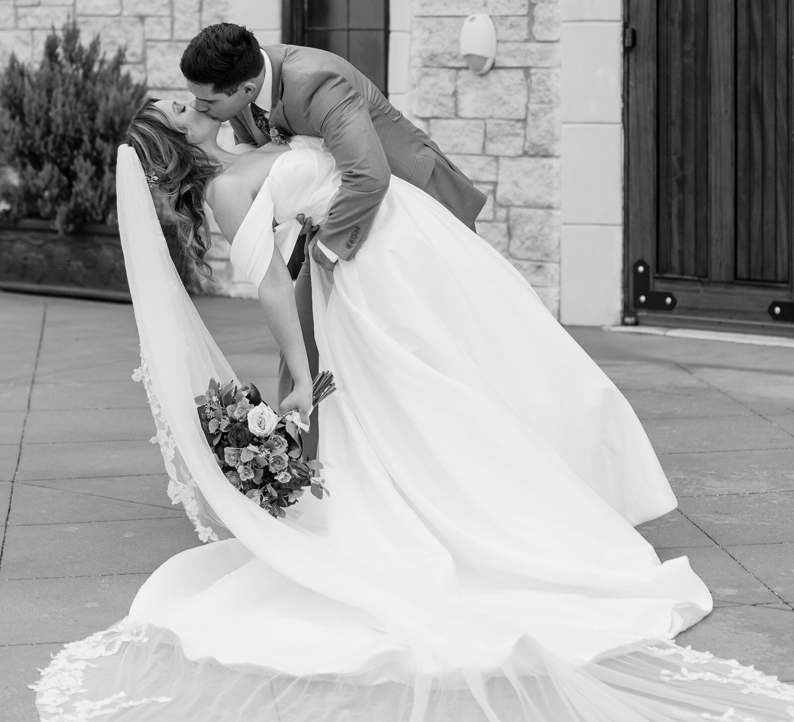 Bride and groom share a romantic dip kiss outside a stone chapel, with the bride’s veil and train fanned elegantly on the ground.
