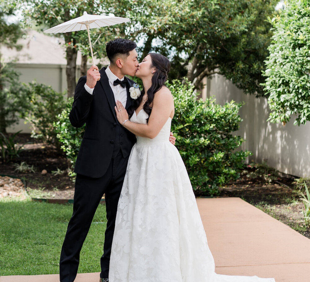 Bride and groom share a kiss outdoors, with the groom holding a white parasol as they stand on a garden pathway surrounded by lush greenery.