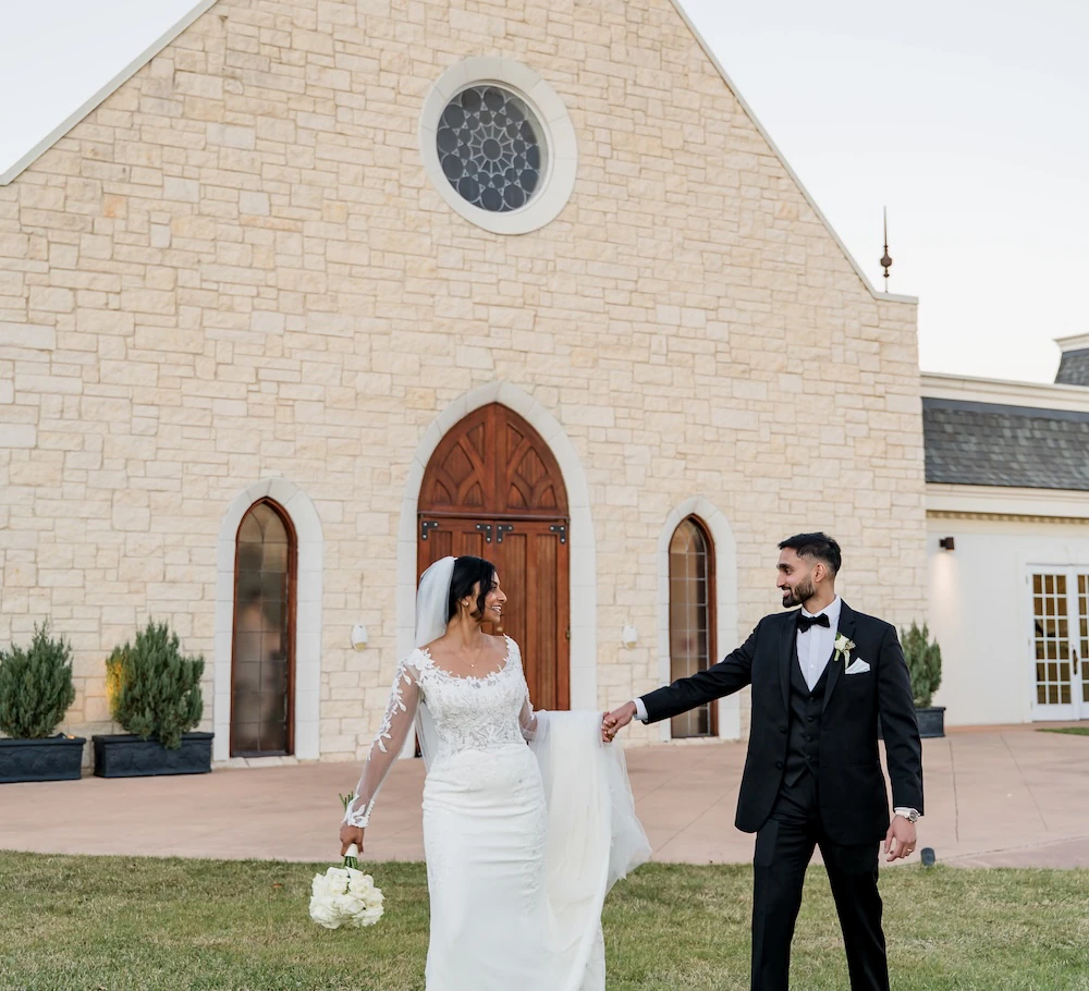 Bride and groom walking hand in hand on the lawn in front of a stone chapel, smiling at each other with the bride holding her bouquet and dress.