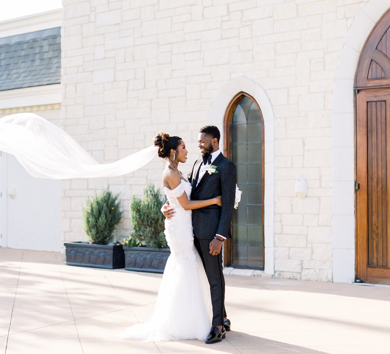 Bride and groom embrace outside a stone chapel as the bride’s veil flows dramatically in the wind behind her.