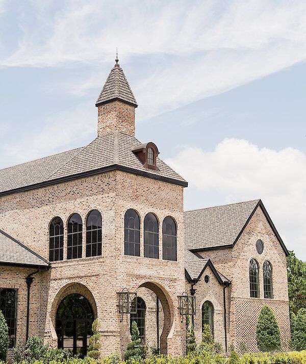 Elegant brick estate-style venue with arched windows, tall tower, manicured landscaping, and a bright blue sky.