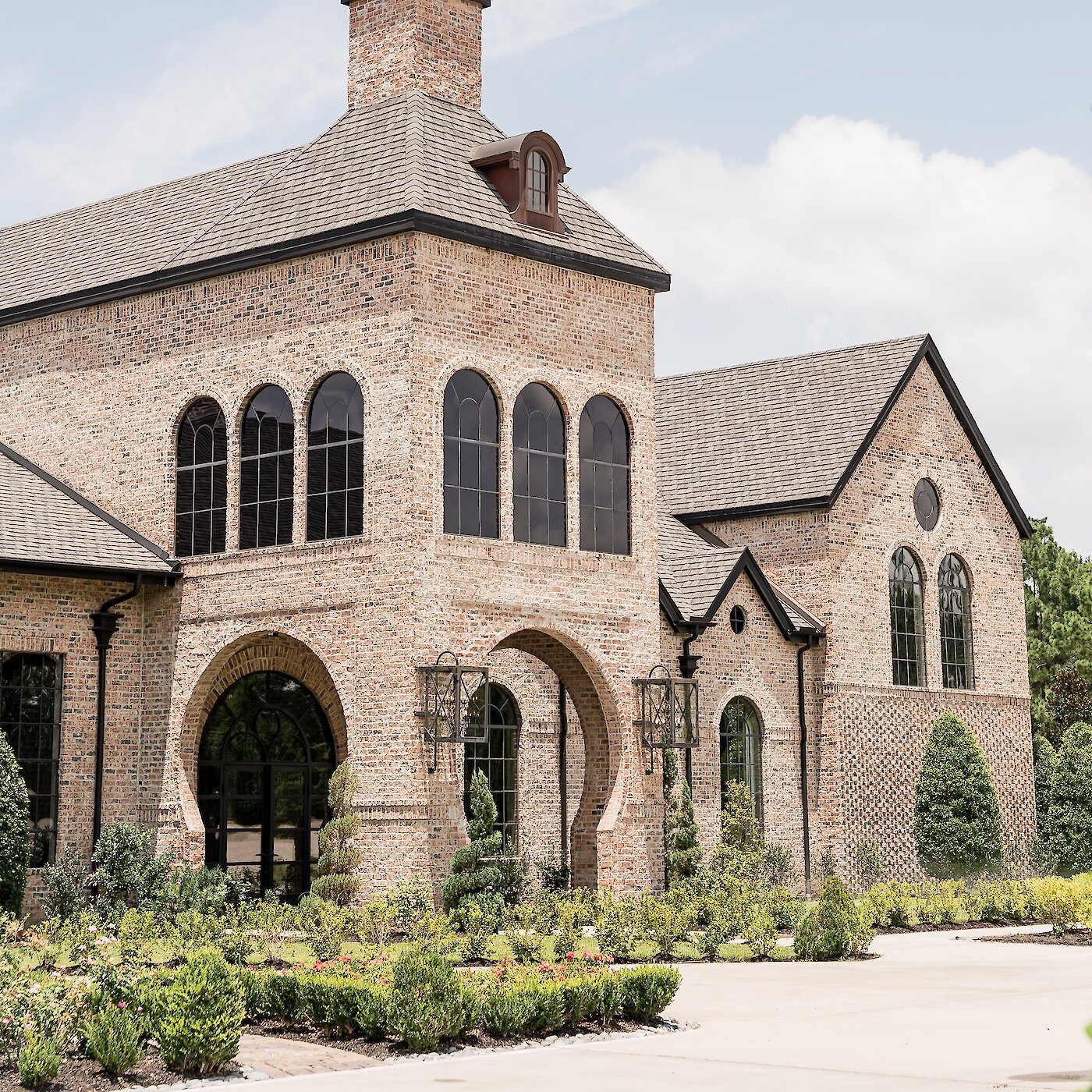 Exterior of an elegant European-style wedding venue constructed with light-colored brick, featuring a steep roof, an entrance tower, and multiple tall arched windows and doorways framed by dark metal.