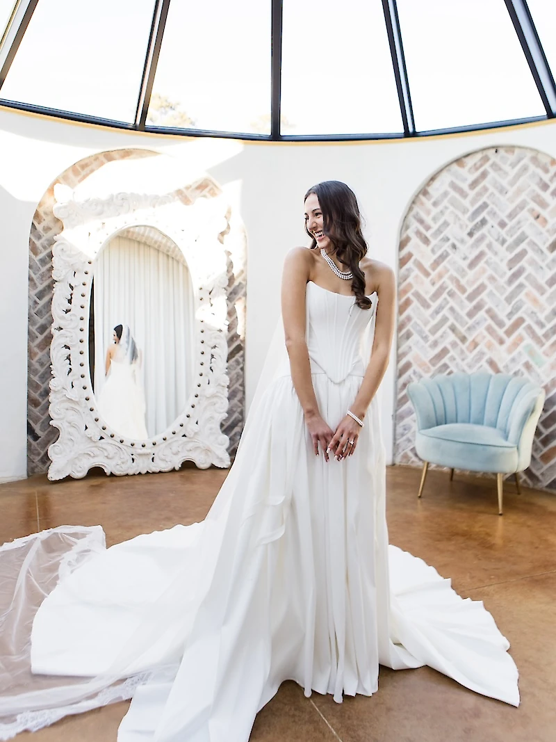 Stunning bridal portrait of the bride in a modern, strapless corset-style white satin gown with an asymmetrical skirt, posing in a suite with a large, ornate white mirror and a pale blue velvet armchair, under a glass dome ceiling.