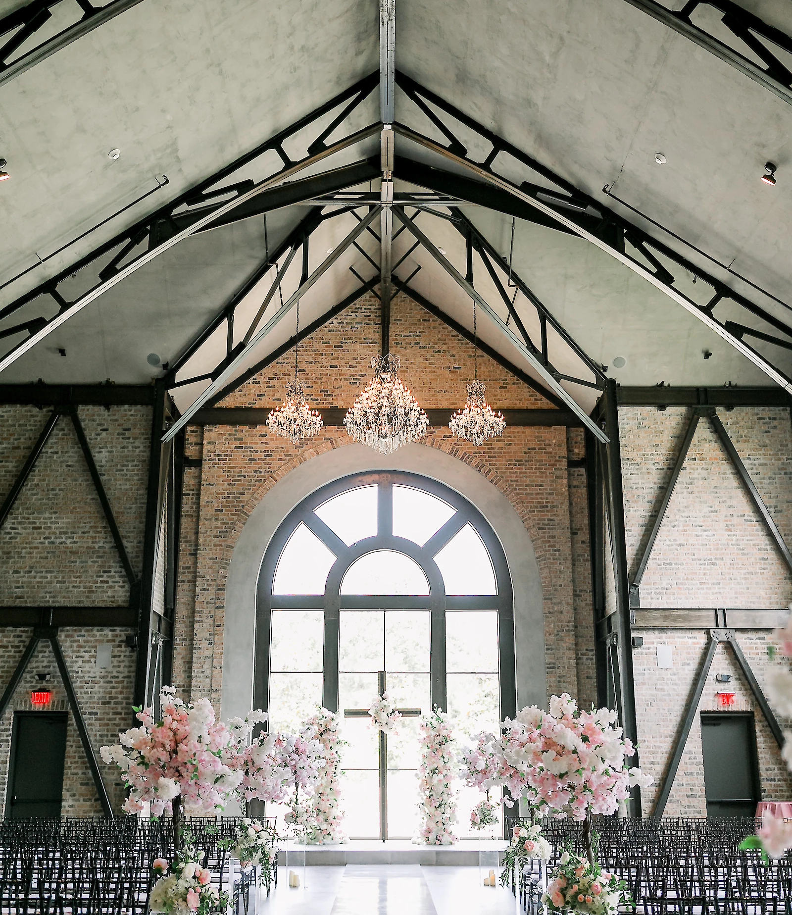 An industrial-style wedding ceremony room with exposed beams and brick, featuring an arched window, crystal chandeliers, and pink floral arrangements lining the aisle.