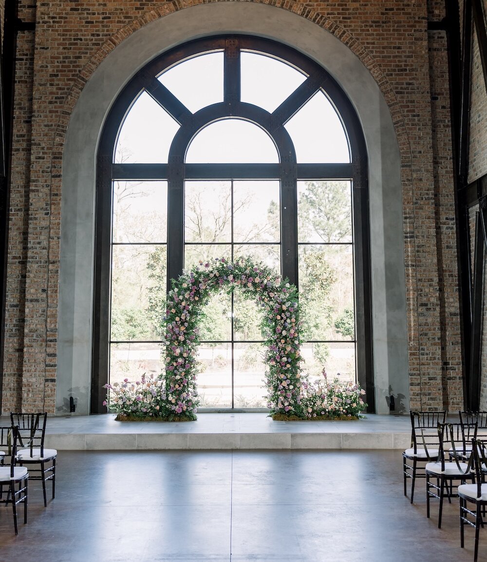 Dramatic wedding ceremony space with a high vaulted ceiling, exposed metal trusses, and brick walls. The altar features a magnificent arched floral ceremony arch in front of a giant arched window, with black Chiavari chairs lining the aisle.