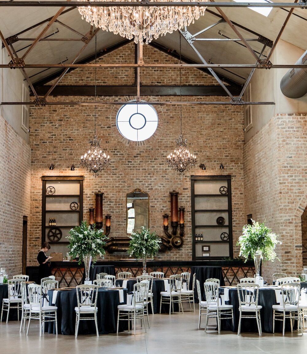 Industrial chic wedding reception hall featuring high vaulted ceilings with exposed metal trusses, rough exposed brick walls, and unique steampunk-style decor. Tables are set with black linens and white chiavari chairs, illuminated by cascading crystal chandeliers.