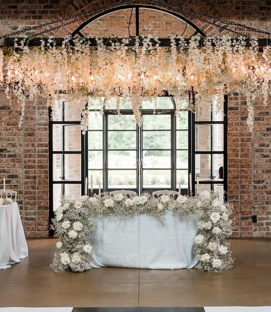 Rustic chic sweetheart table set against an exposed brick wall and arched window. The table is draped with white linens and surrounded by a lavish garland of white baby's breath and roses. A dramatic hanging floral chandelier is suspended above.