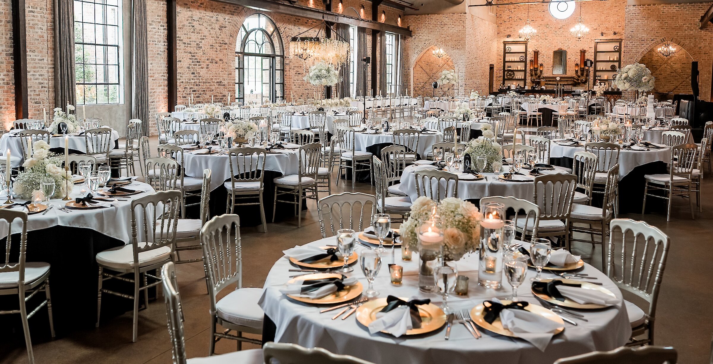 An industrial-chic wedding reception setup with brick walls, high ceilings, large windows, and round tables set with gold chargers and white floral centerpieces.