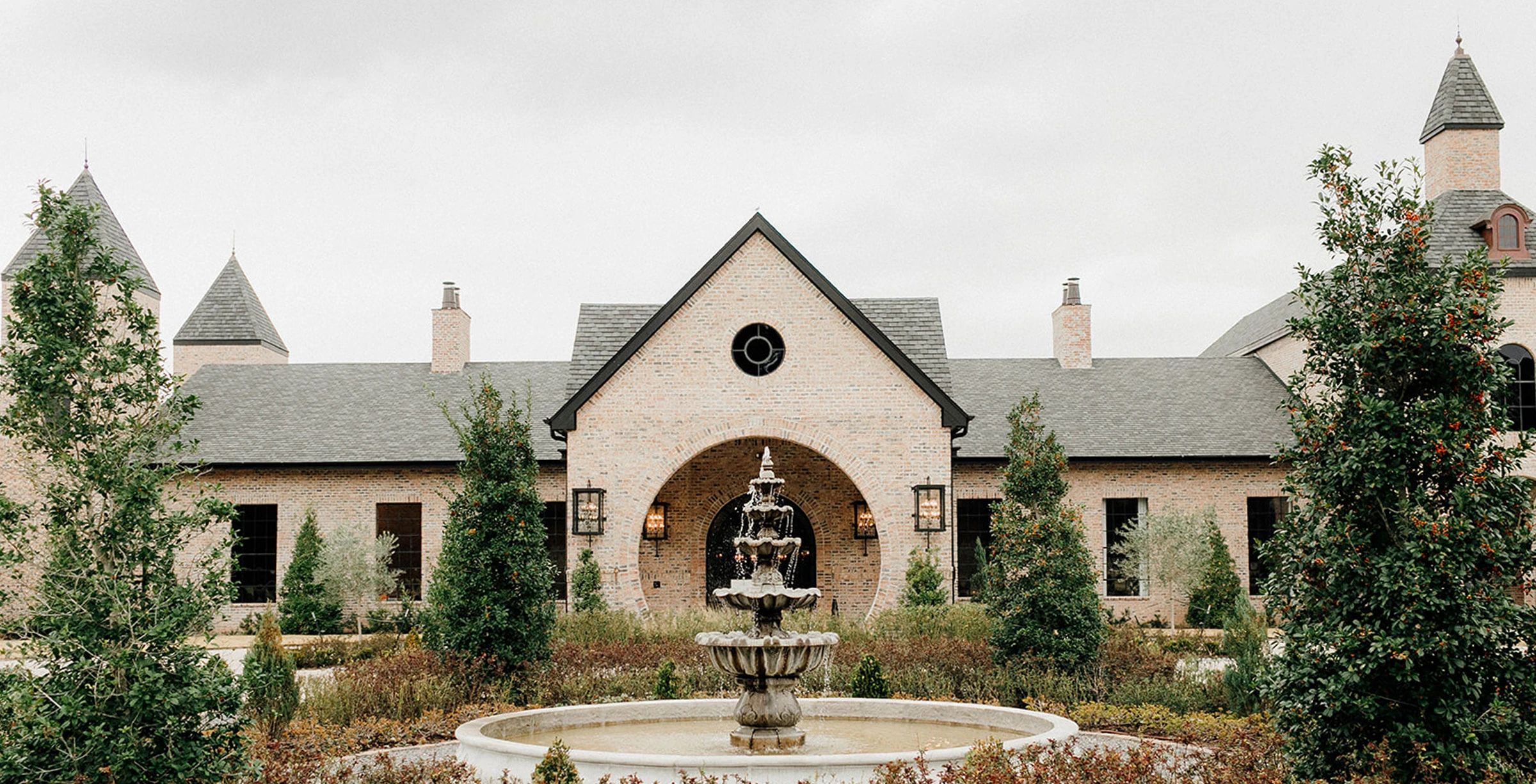 Elegant wedding portrait of the couple posing at the edge of a tiered stone fountain in a manicured garden. The groom wears a black tuxedo and the bride wears a fitted lace gown with a flowing veil, holding a lush bouquet of purple, pink, and white flowers.