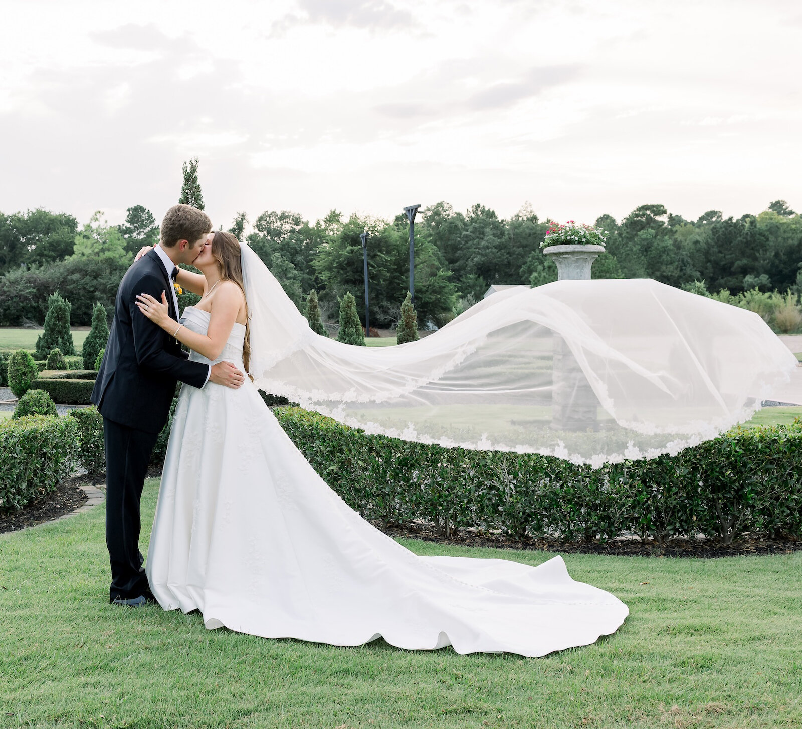 Romantic outdoor wedding kiss with the bride's long cathedral veil flowing dramatically in the wind. The couple is embracing on a manicured green lawn with trimmed hedges and sculpted topiaries in the background.