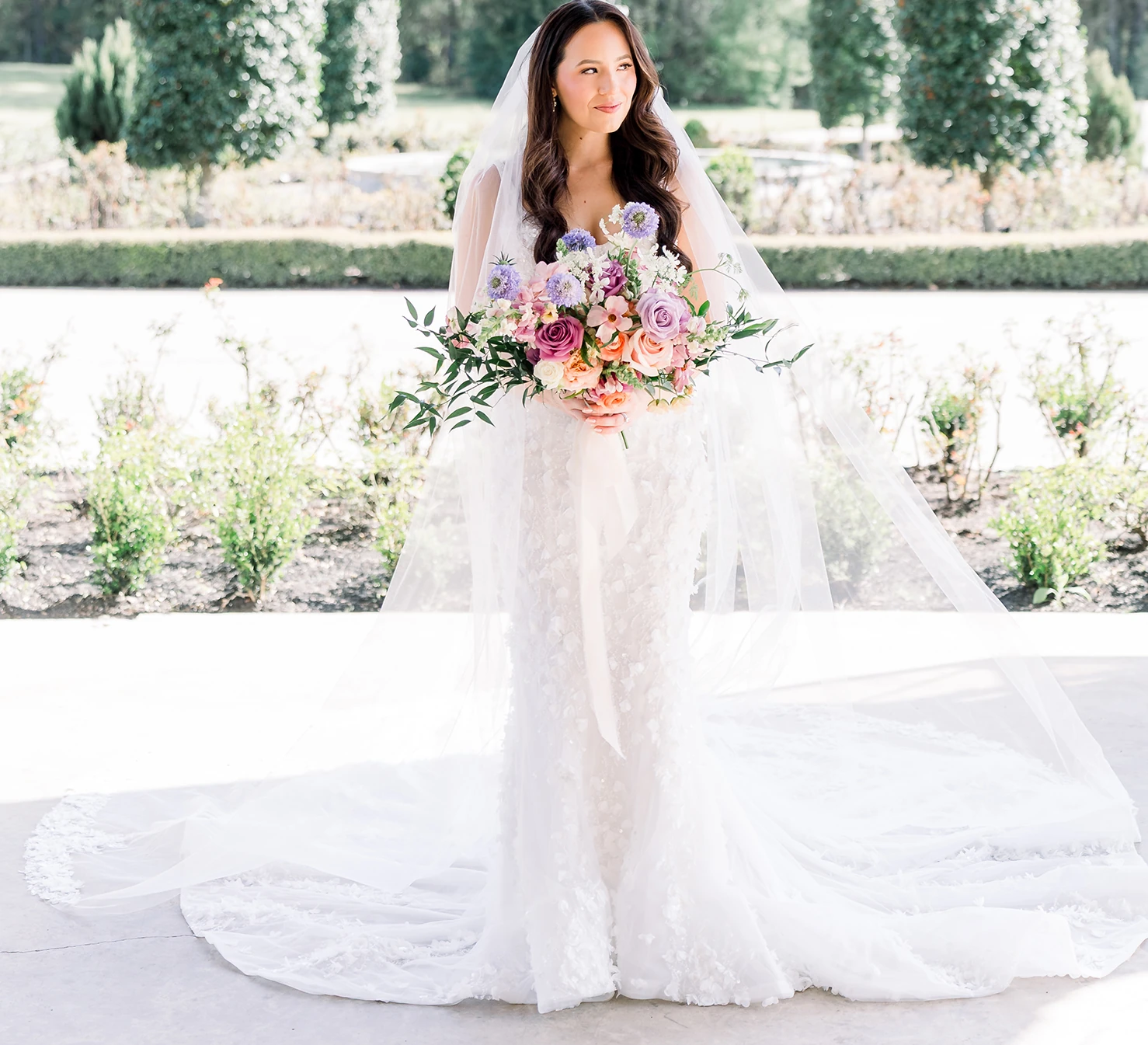 Bride in a textured lace gown and veil holding a vibrant bouquet of lavender, pink, and peach pastel flowers in a garden setting