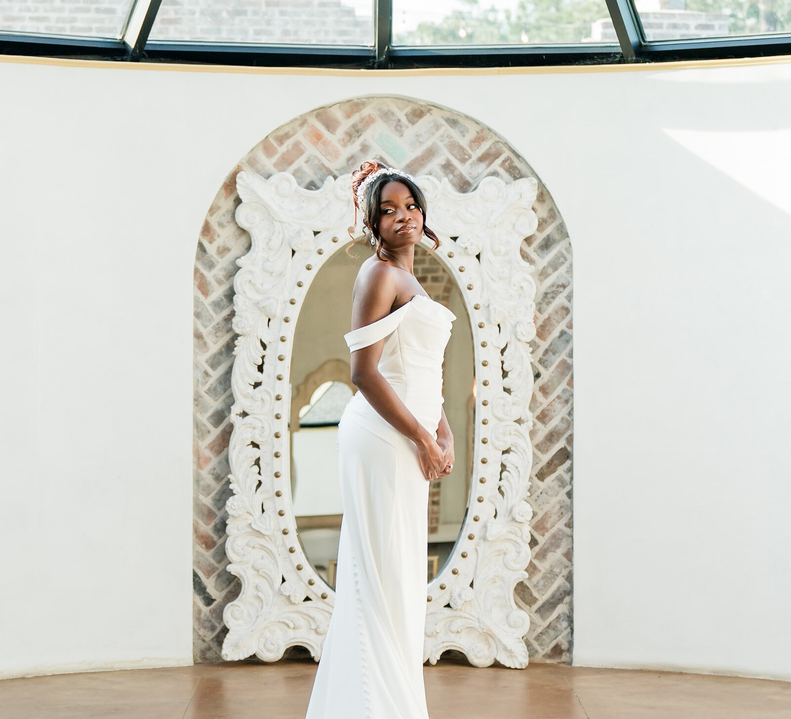A beautiful bride in a sleek, off-the-shoulder white wedding dress stands on a round rug under a glass dome ceiling with a chandelier.
