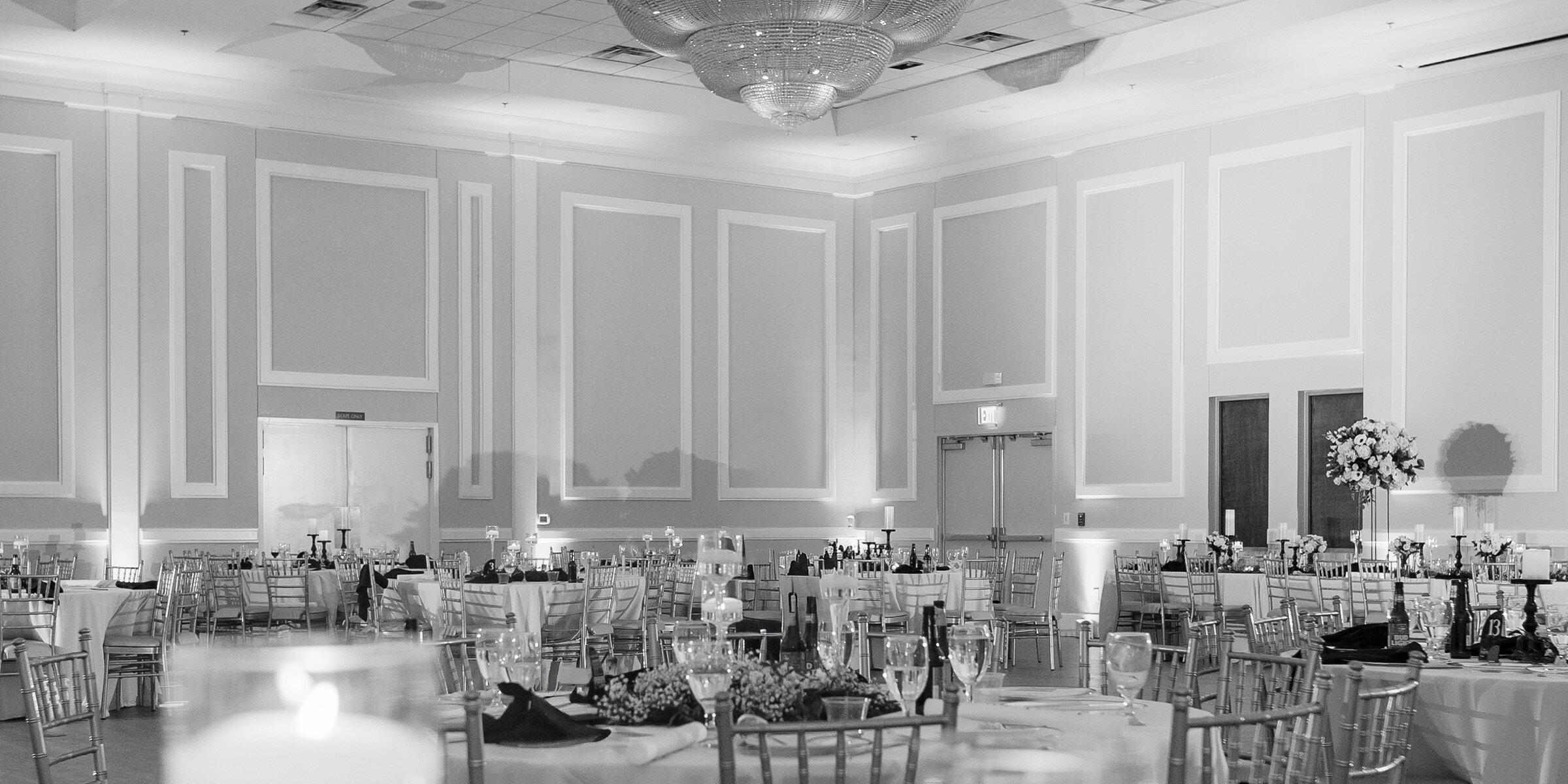 Dramatic industrial wedding reception hall featuring high ceilings, rough exposed brick walls, and arched doorways. Tables are set with white linens, white Chiavari chairs, and lavish deep red floral centerpieces, beneath ornate chandeliers and unique industrial-style steampunk wall decor.