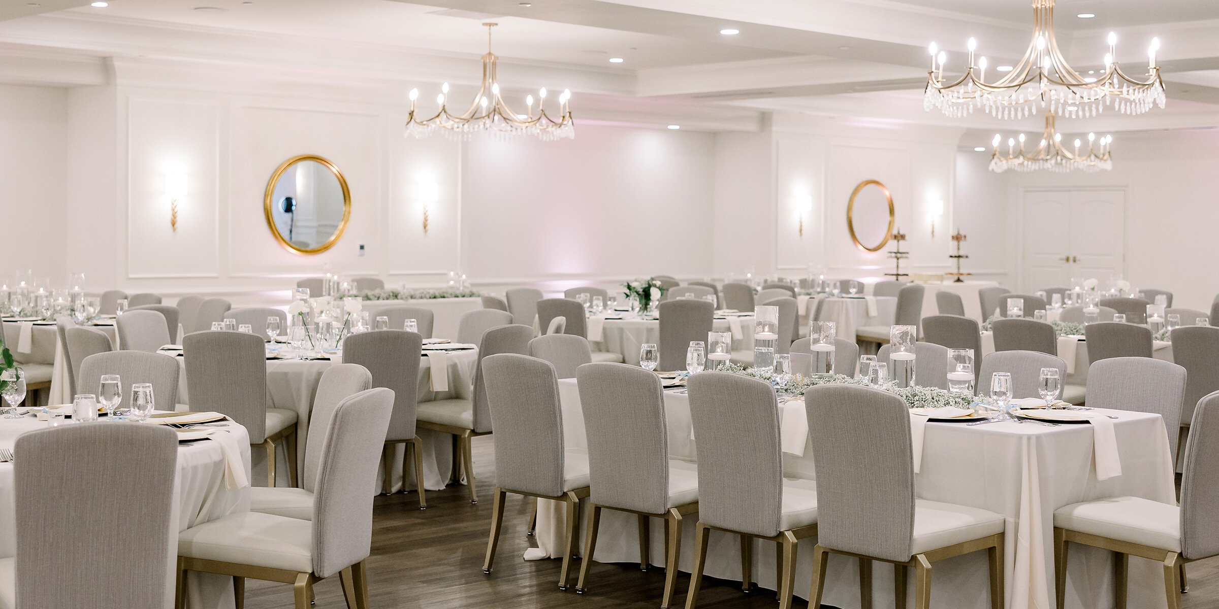 A luxurious, bright ballroom set for a reception with long rectangular tables covered in blush linen, surrounded by light grey chairs and gold chandeliers.