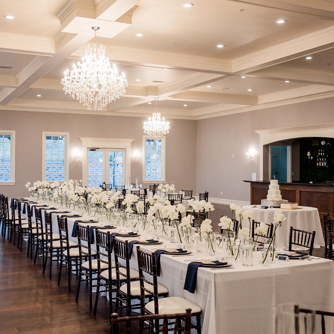 Warm rustic wedding reception interior inside a log cabin-style venue with wood-paneled walls, exposed beams, and string bistro lights. Round tables are set with textured white linens, wooden folding chairs, and simple white floral centerpieces.