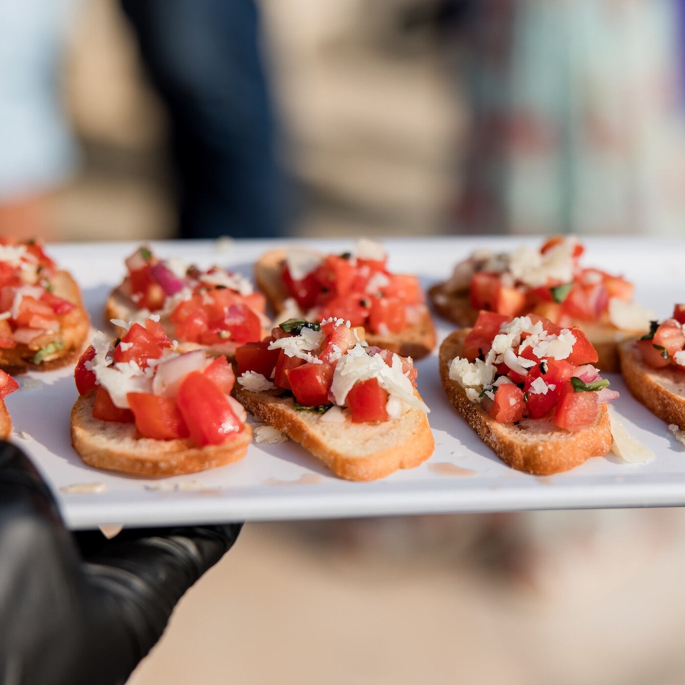A server wearing a black glove holds a white tray of fresh bruschetta appetizers topped with diced tomatoes and grated cheese.