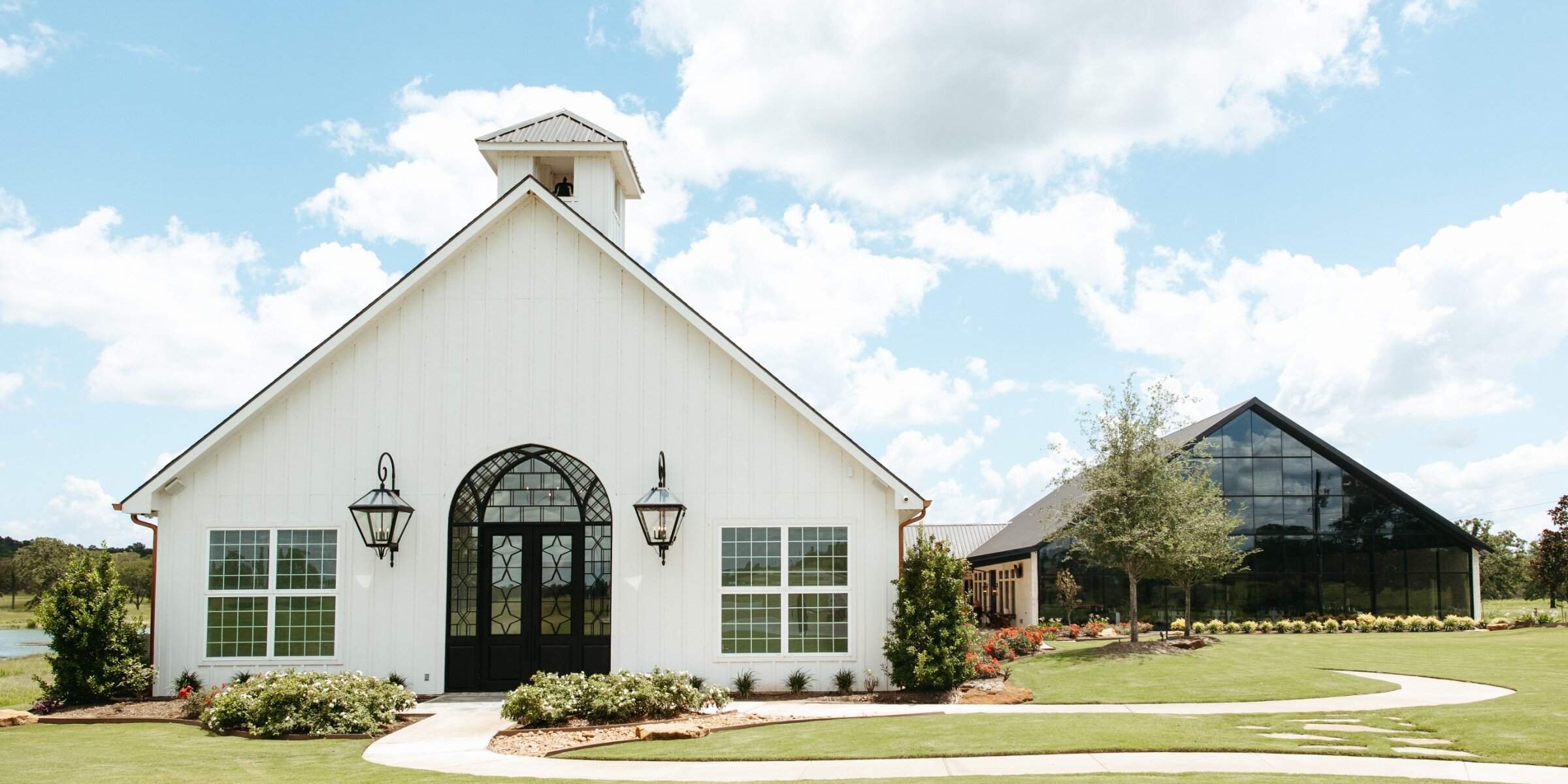 Exterior of a modern white chapel wedding venue with vertical siding, a central arched black-framed door, and a small bell tower. The complex is set on a manicured green lawn under a blue sky, next to a contemporary glass building structure.
