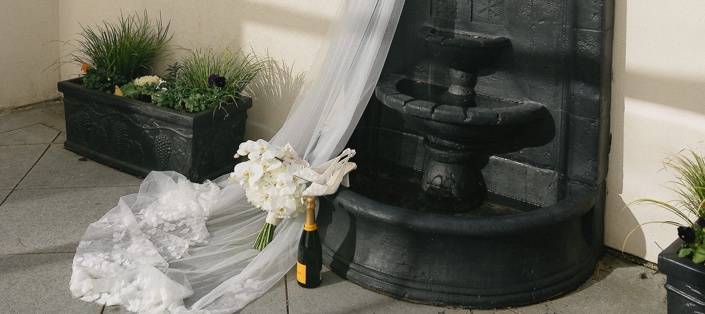 Black wall fountain with wedding veil draped over it with heels, bouquet and champagne artfully placed