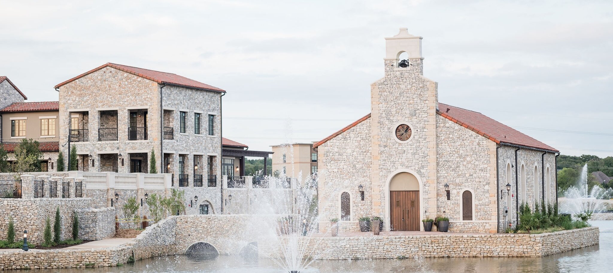 Wide exterior view of a Mediterranean-style wedding venue complex featuring a stone chapel with a bell tower and a multi-story reception building, all situated on a pond with a central water fountain.
