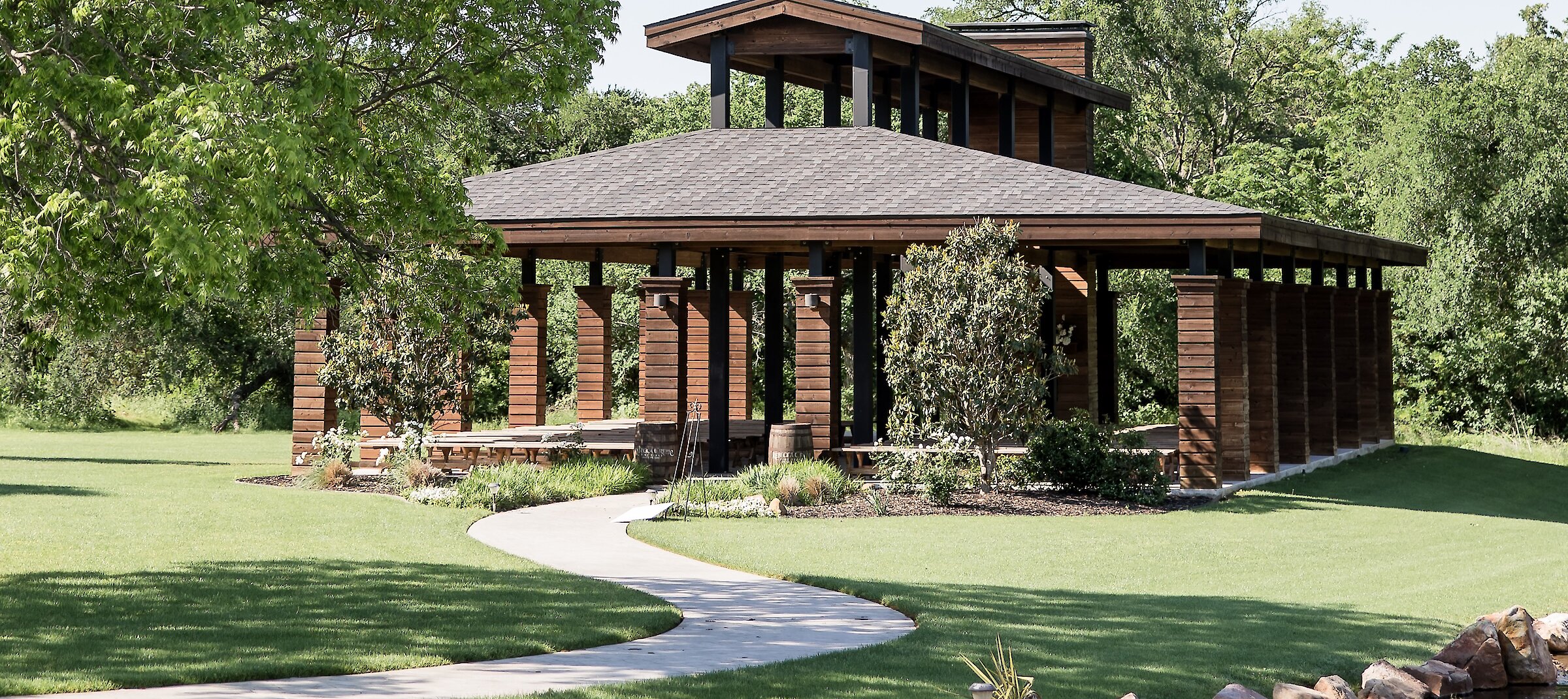A Frank Lloyd Wright-inspired outdoor ceremony pavilion with a dark wood structure sits on a green lawn with a winding path and small creek.