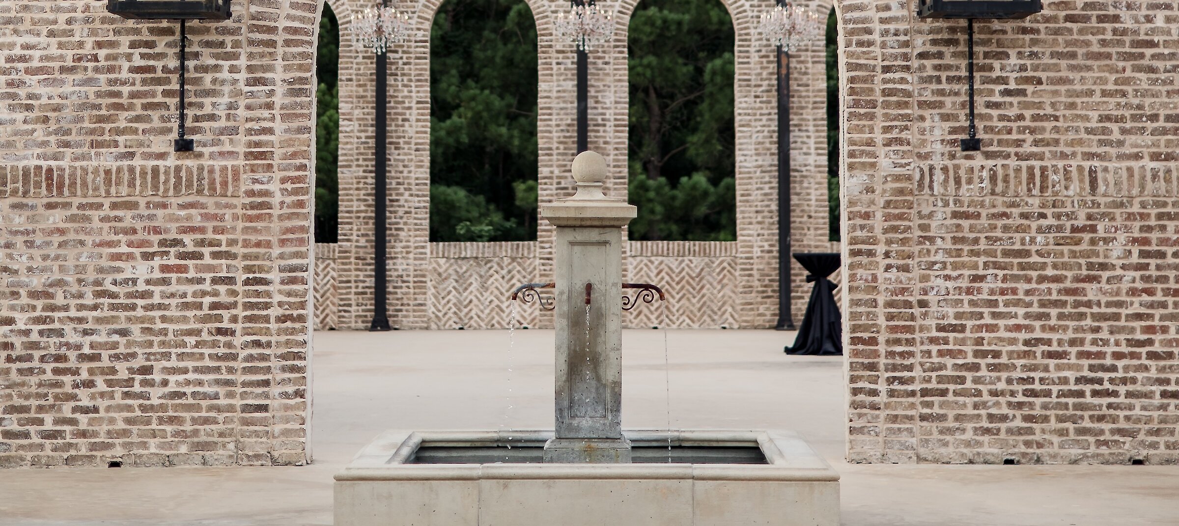 An outdoor ceremony space featuring an arched brick ruin wall with hanging lanterns and a small stone fountain in the foreground.