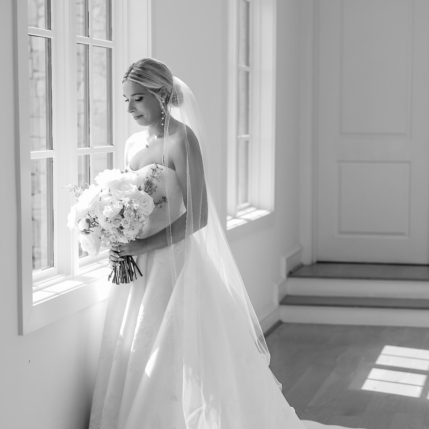 Black and white photo of bride standing in a sunny window looking at her bouquet