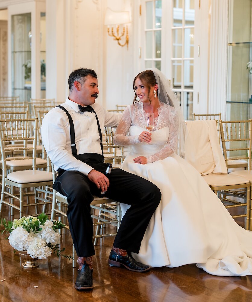Bride with champagne and groom with beer sit together in empty hall sharing a smile after wedding ceremony