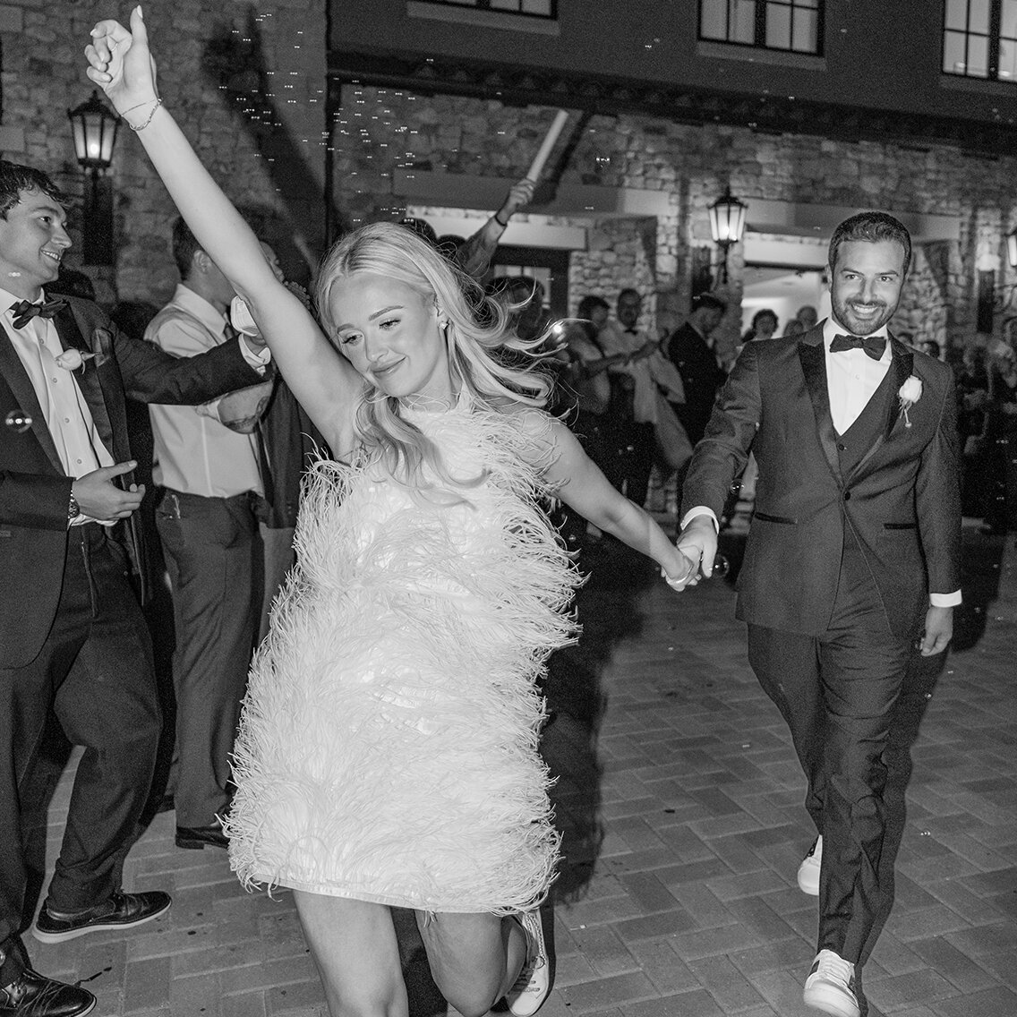 Black and white photo of the bride and groom during their grand exit, hand-in-hand. The bride is running and celebrating in a short feathered reception dress and sneakers, while the groom smiles, both surrounded by guests outside a stone venue.