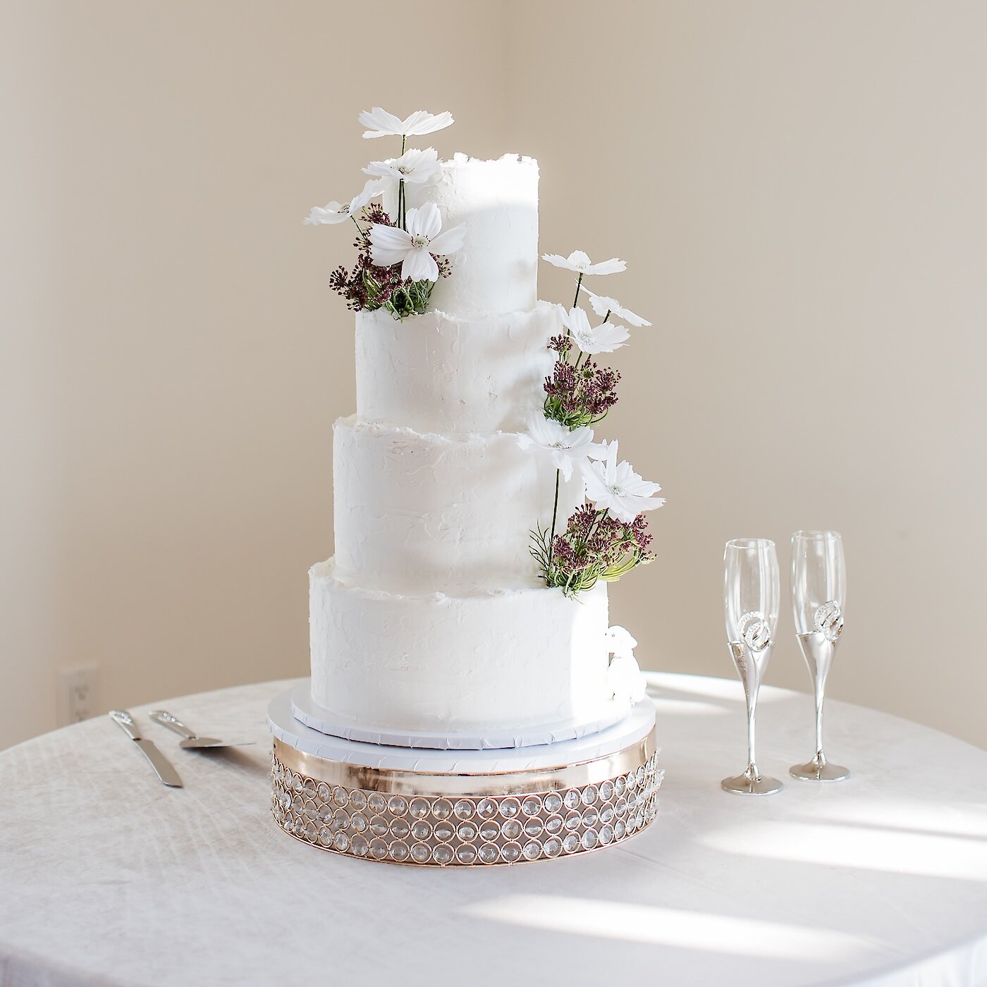 A four-tiered white textured wedding cake decorated with white cosmos flowers and greenery, sitting on a gold pedestal stand next to two champagne flutes.